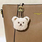 A close-up of a tan pebbled leather handbag with a small white fuzzy teddy bear keychain charm hanging from a gold-tone ring and strap.