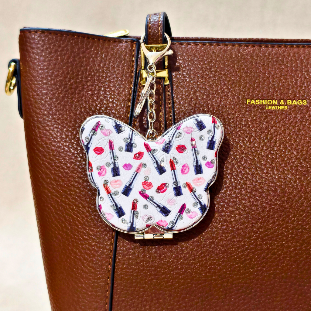 A close-up of a brown pebbled leather handbag with a butterfly-shaped charm hanging from the zipper. The charm has a white background with a colorful pattern of lipstick bullets and red lips, and is attached with gold-tone hardware.