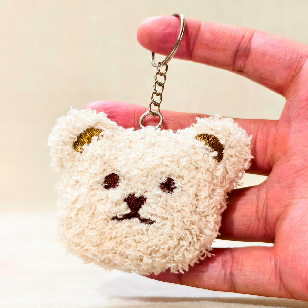 A small plush bear-face keychain held in a person's hand. The keychain is cream-colored with embroidered brown facial features and gold-toned ear accents, attached to a silver metal ring and chain.