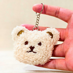 A small plush bear-face keychain held in a person's hand. The keychain is cream-colored with embroidered brown facial features and gold-toned ear accents, attached to a silver metal ring and chain.