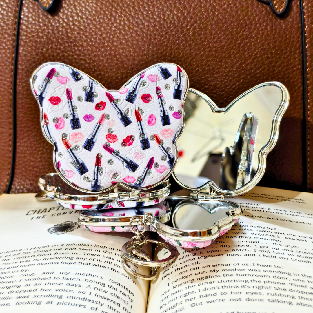 A butterfly-shaped compact mirror with a white patterned top half featuring lipstick tubes and lip prints, and a mirrored bottom half, displayed open on an open book with a brown leather bag in the background.