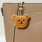 A close-up of a tan textured leather handbag with a small fuzzy teddy bear keychain charm hanging from the zipper pull.