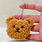 A plush keychain shaped like a cartoon Teddy face, featuring embroidered black eyes, and mouth details, a small nose, and a silver metal key ring and chain On a Beige Background Shown hand carry.