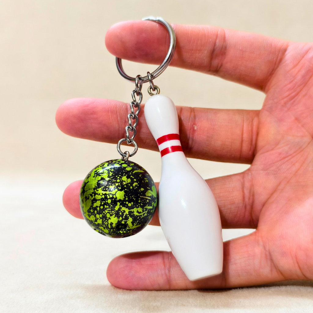 A novelty bowling keychain held in a hand, featuring a green and black mottled bowling ball and a white bowling pin with a red stripe, attached to a silver split ring.
