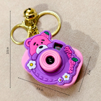 A pink and purple cartoon camera keychain with a gold bell and key ring, shown on a beige fabric background with dimension lines indicating measurements of 3.5 in, 2.