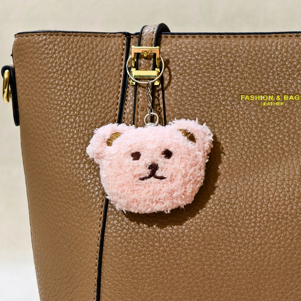 A light pink fluffy teddy bear head keychain charm with gold-tone ears and black embroidered facial features, hanging from a gold clasp on a brown textured leather handbag.