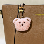A light pink fluffy teddy bear head keychain charm with gold-tone ears and black embroidered facial features, hanging from a gold clasp on a brown textured leather handbag.
