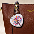 A heart-shaped silver metal compact case with a colorful butterfly tree design, attached to a brown textured leather handbag.