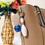 A close-up of a tan textured handbag with a metal key ring and two hanging charms: a white bowling pin with red stripes and a blue marbled bowling ball. A decorative vase with flowers sits in the blurred background.