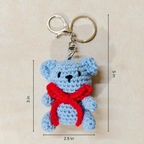 Crocheted teddy bear keychain with a red bow, shown with measurements on a beige background.