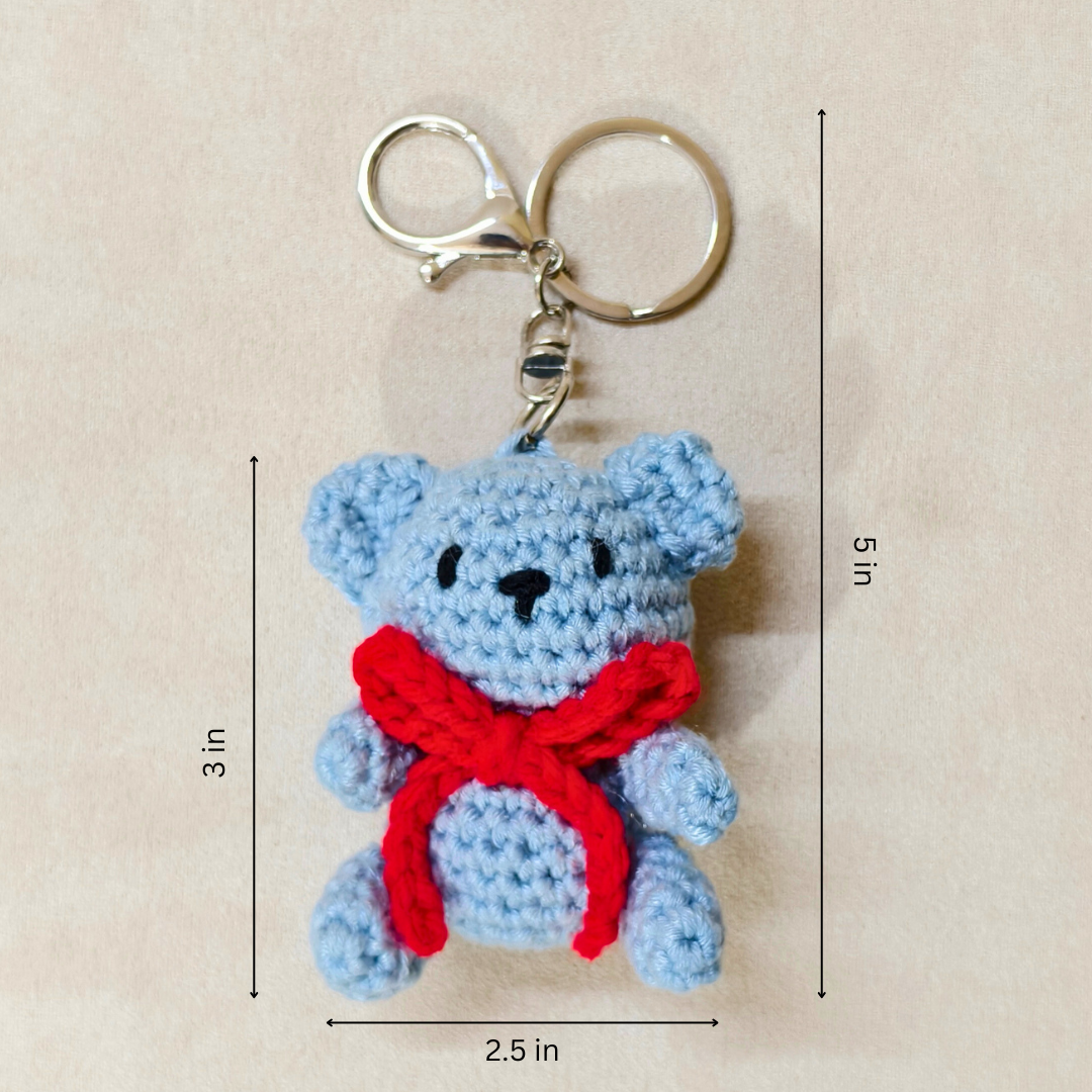 Crocheted teddy bear keychain with a red bow, shown with measurements on a beige background.