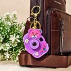 A purple and pink cartoon bear camera keychain charm with white daisy accents and a gold key ring, attached to a brown leather bag with a brown shoulder strap, set against a background of faux green and white flowers.