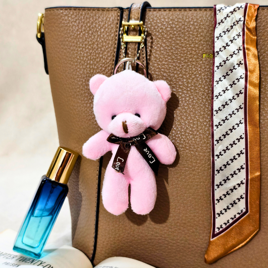 A small pink plush teddy bear keychain charm with a black ribbon bow reading 'Love' hangs from a brown textured handbag. The bag has a gold zipper and a white patterned scarf draped over the side.