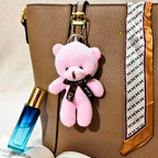 A small pink plush teddy bear keychain charm with a black ribbon bow reading 'Love' hangs from a brown textured handbag. The bag has a gold zipper and a white patterned scarf draped over the side.