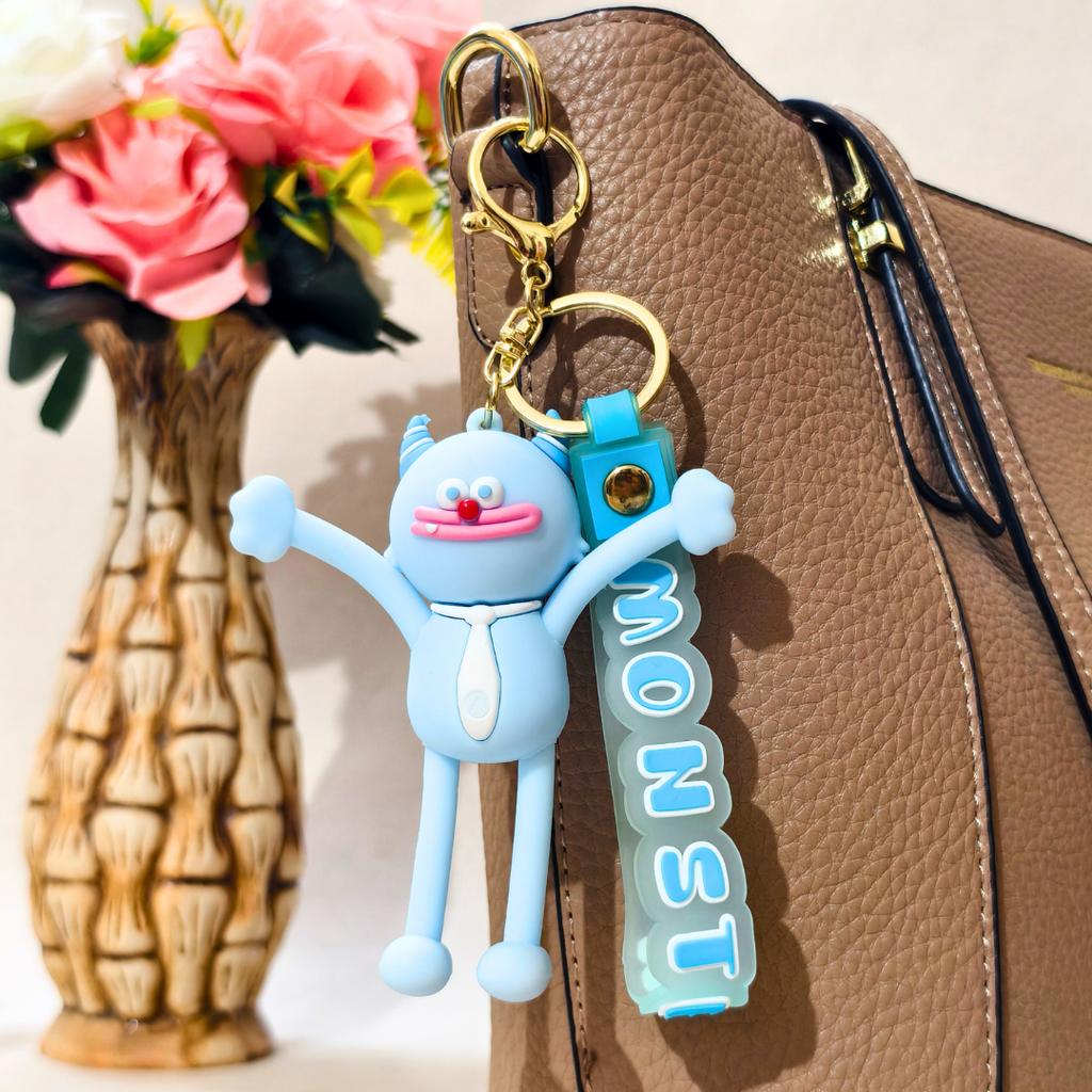 A light blue 3D cartoon keychain charm with arms and a matching blue strap reading 'MONST' hangs from gold hardware next to a brown textured handbag, with a vase of pink flowers blurred in the background.