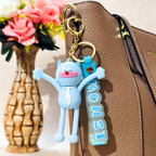 A light blue 3D cartoon keychain charm with arms and a matching blue strap reading 'MONST' hangs from gold hardware next to a brown textured handbag, with a vase of pink flowers blurred in the background.