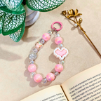 Pink beaded keychain with heart charm on a book next to green leaves and gold flower