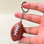 A miniature brown American football keychain with white laces and a silver split ring, held between fingers to show its small scale.