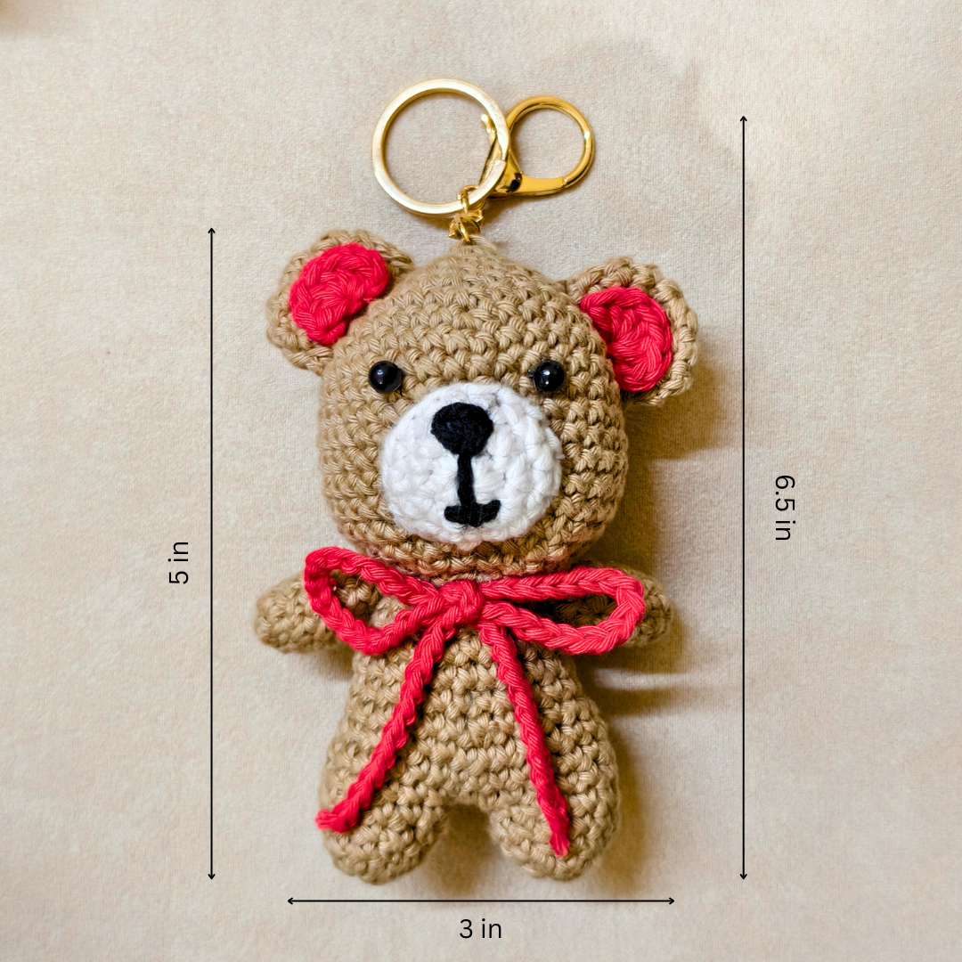 Crocheted teddy bear keychain with red bow and ears, shown with measurements on a beige background.