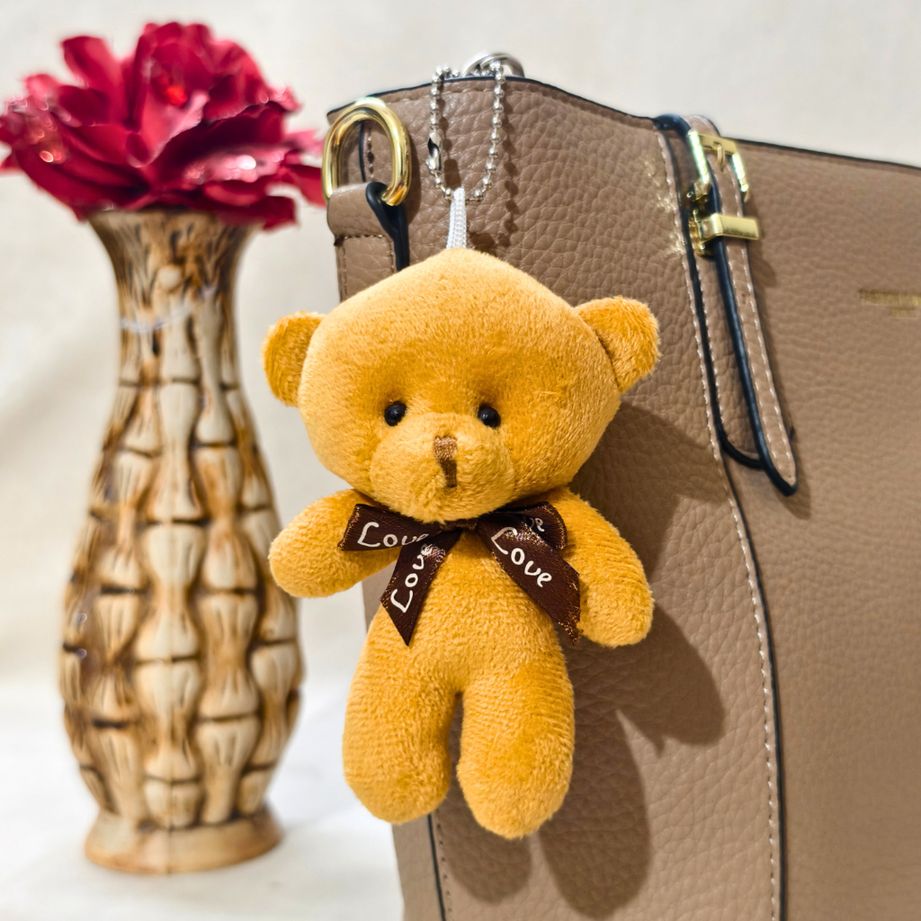Teddy bear keychain with 'Love' bow tie attached to a brown leather bag, with a red flower in the background.