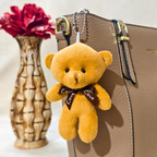 Teddy bear keychain with 'Love' bow tie attached to a brown leather bag, with a red flower in the background.