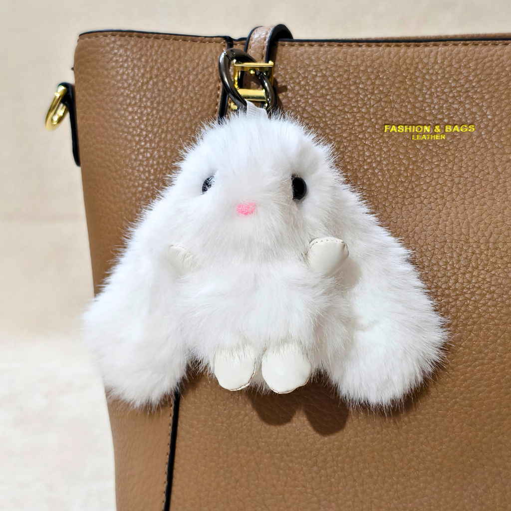 A fluffy white faux-fur bunny keychain charm with black eyes and a pink nose, hanging from a gold ring on a brown textured handbag.