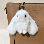 A fluffy white faux-fur bunny keychain charm with black eyes and a pink nose, hanging from a gold ring on a brown textured handbag.