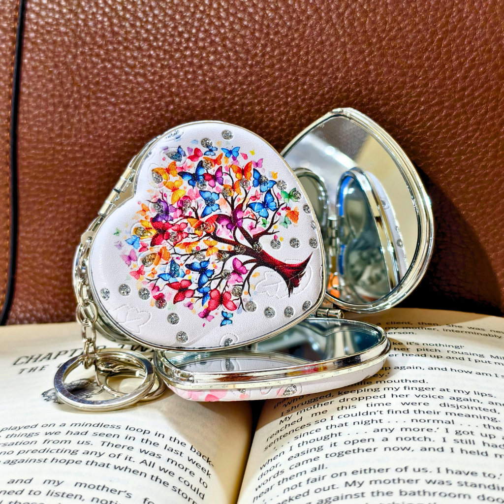 A heart-shaped silver keepsake box with a colorful butterfly tree of life design, resting on an open book against a brown textured leather background.