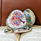 A heart-shaped silver keepsake box with a colorful butterfly tree of life design, resting on an open book against a brown textured leather background.