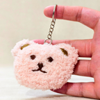 A small plush bear-shaped keychain in pale pink with embroidered brown facial features and gold ear accents, held between a person's fingers against a soft beige background.