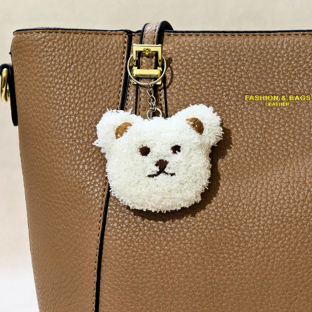 A white fluffy teddy bear face charm with brown ears, nose, and eyes, hanging from a gold-tone ring on a brown textured leather bag.