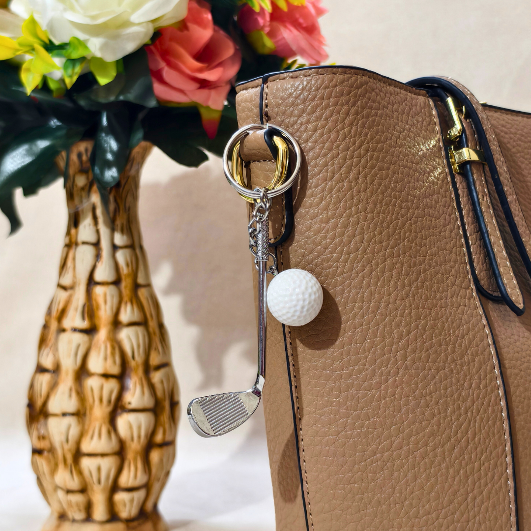 A tan pebbled leather handbag with a silver-tone key ring and a golf-themed keychain charm featuring a white golf ball and a miniature golf club head, shown next to a decorative ceramic vase with flowers.