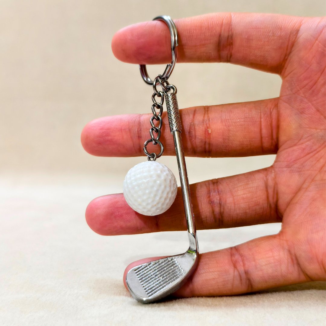 A hand holding a miniature golf keychain featuring a silver metal golf club head and a white textured golf ball charm, set against a neutral beige background.