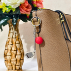 A silver metal keychain featuring a miniature golf club head and a bright red textured pom-pom ball, attached to a tan pebbled leather handbag. A decorative ceramic vase with flowers is visible in the background.