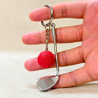 A miniature silver metal golf club keychain with a textured red golf ball charm, held between a person's fingers against a neutral beige background.
