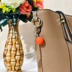 A miniature golf club keychain with an orange textured golf ball charm, hanging on Handbag as Bag Charm against a neutral beige background.