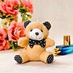 A small plush teddy bear toy in tan with a white snout, black polka-dot bow tie, and black polka-dot ear and paw pads, posed on a light beige surface with pink flowers and a blue perfume bottle in the background.