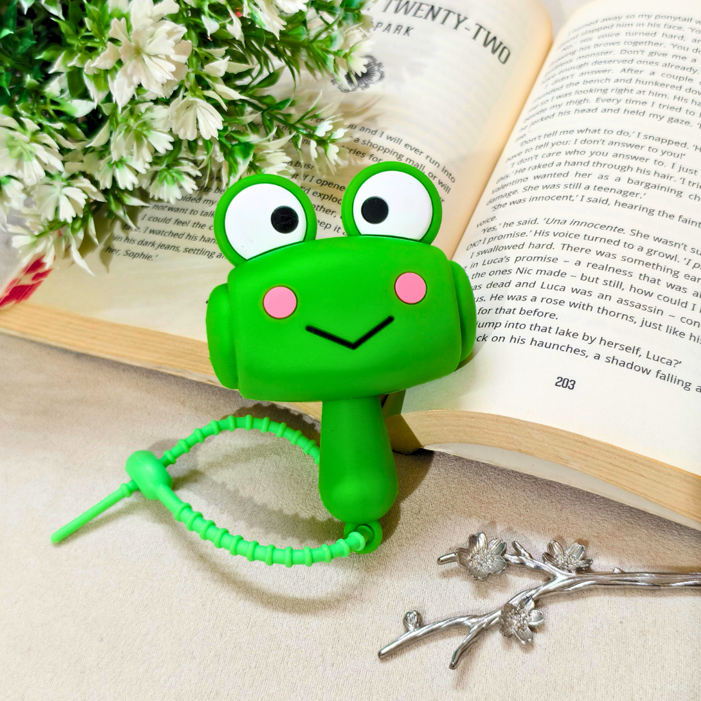 A green frog-shaped book end with large cartoon eyes, pink cheeks, and a coiled tail, displayed on an open book with decorative white flowers and silver branches.