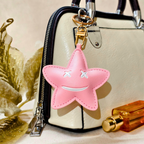 Pink star-shaped keychain attached to a handbag with a gold flower and perfume bottle in the background.
