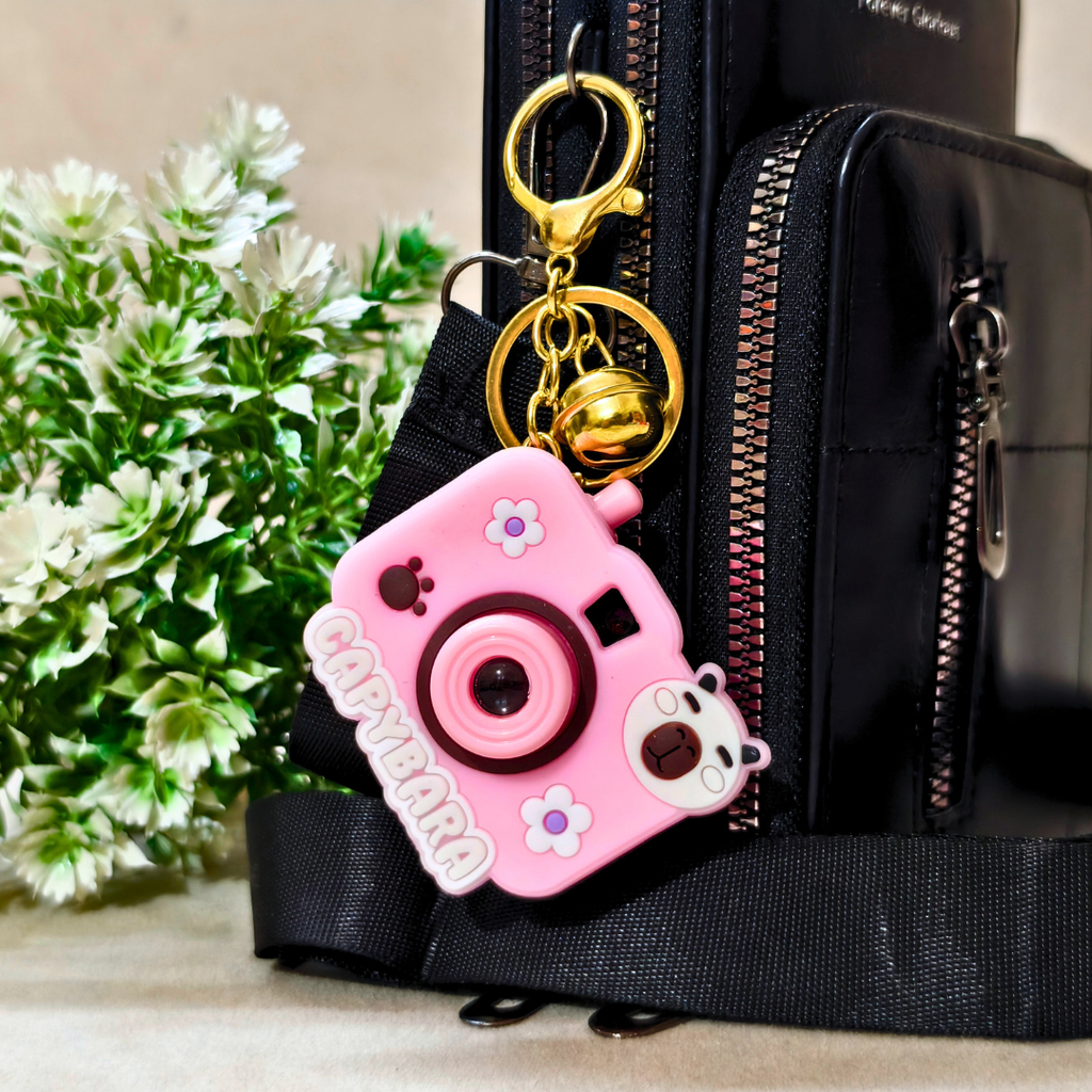 Pink camera-shaped keychain attached to a black bag with a plant in the background