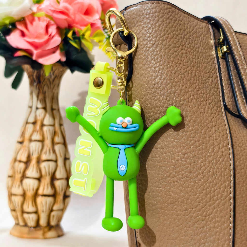 A green cartoon monster keychain charm with a translucent neon yellow name tag reading 'ISNWA' and a gold key ring, shown hanging beside a brown textured handbag with pink flowers in a vase in the background.