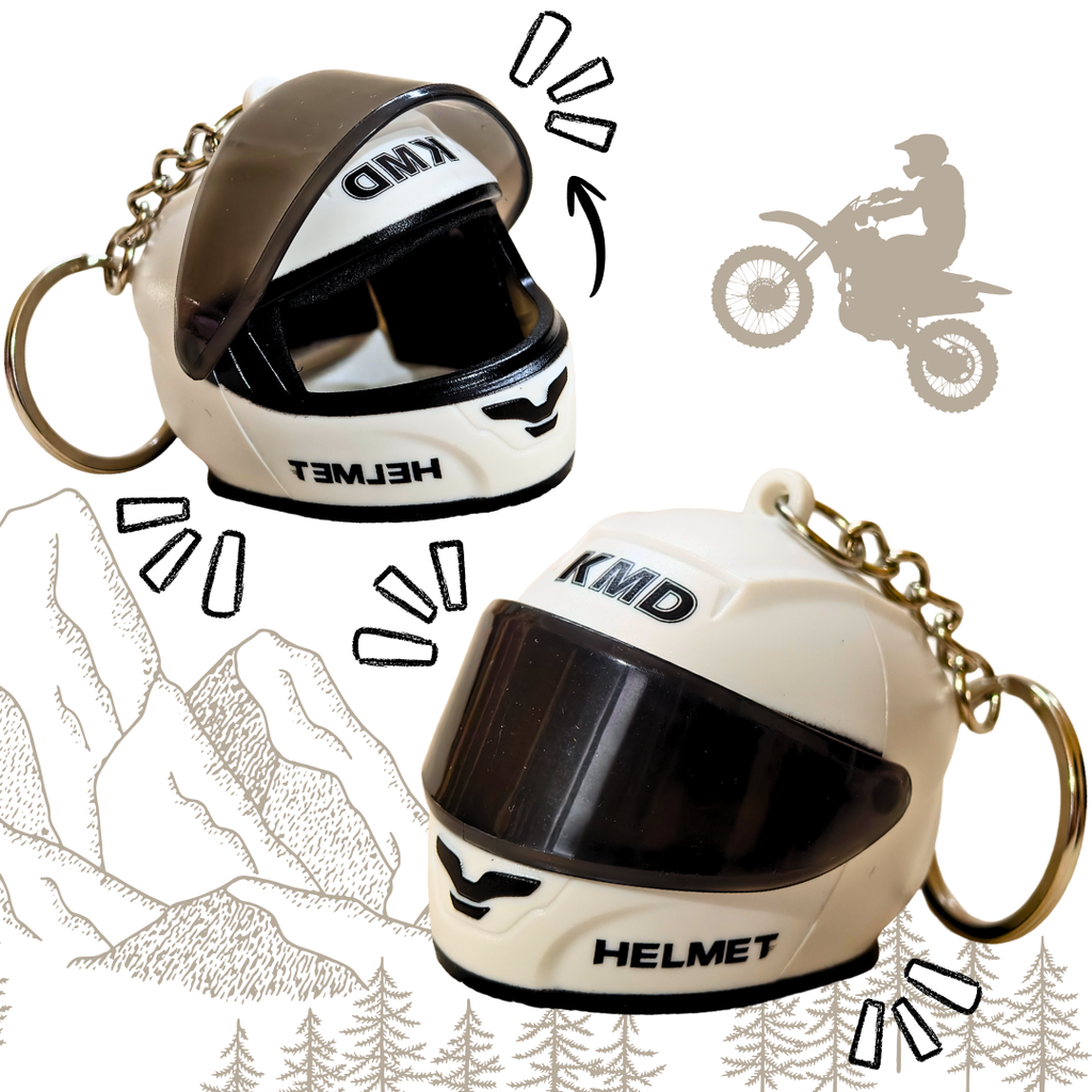 Two small motorcycle helmet keychains shown on a white background with decorative line drawings of mountains, trees, and a motorcycle silhouette. The keychains are cream/off-white with black visors and black printed text reading 'KMD' and 'HELMET'; one also has 'T3MID' visible on the forehead area.