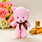 A small pink plush teddy bear standing upright, wearing a dark brown ribbon with 'Love' printed on it. The bear is styled on a light beige surface with a gold perfume bottle on the right and a bouquet of pink and white roses in the background.