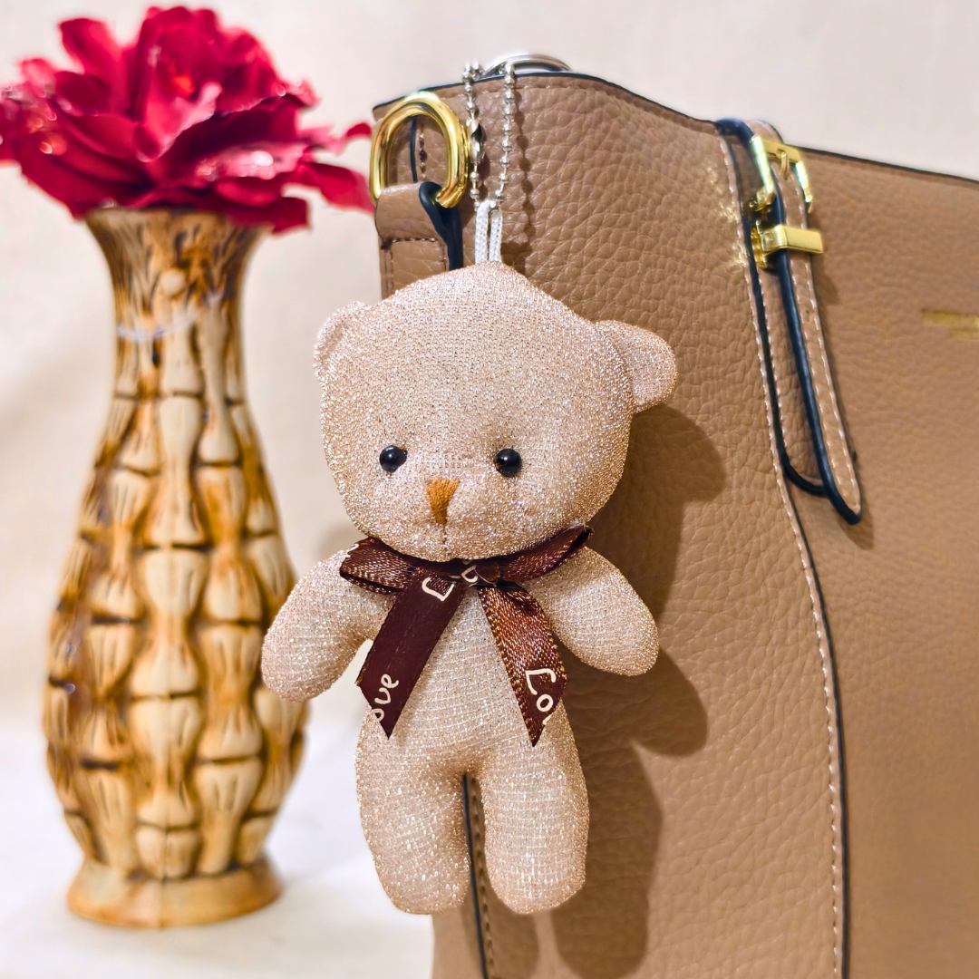 Teddy bear keychain with 'Love' ribbon attached to a beige handbag, with a vase of red flowers in the background.