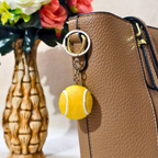 A yellow tennis ball keychain charm with white lines and a silver metal key ring, attached to a brown textured leather handbag. A decorative vase with flowers is blurred in the background.