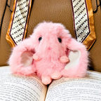 A fluffy pink plush bunny toy with long ears rests on open book pages, with patterned neckties in the background.