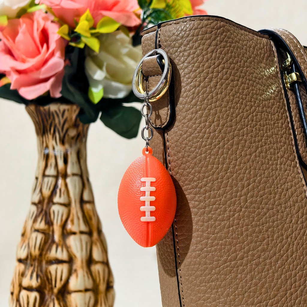 A bright orange football-shaped keychain with white laces hanging from a silver key ring, shown attached to a brown textured handbag next to a decorative vase with pink flowers.