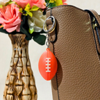 A bright orange football-shaped keychain with white laces hanging from a silver key ring, shown attached to a brown textured handbag next to a decorative vase with pink flowers.
