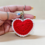 Hand holding a red heart-shaped crochet keychain on a beige background