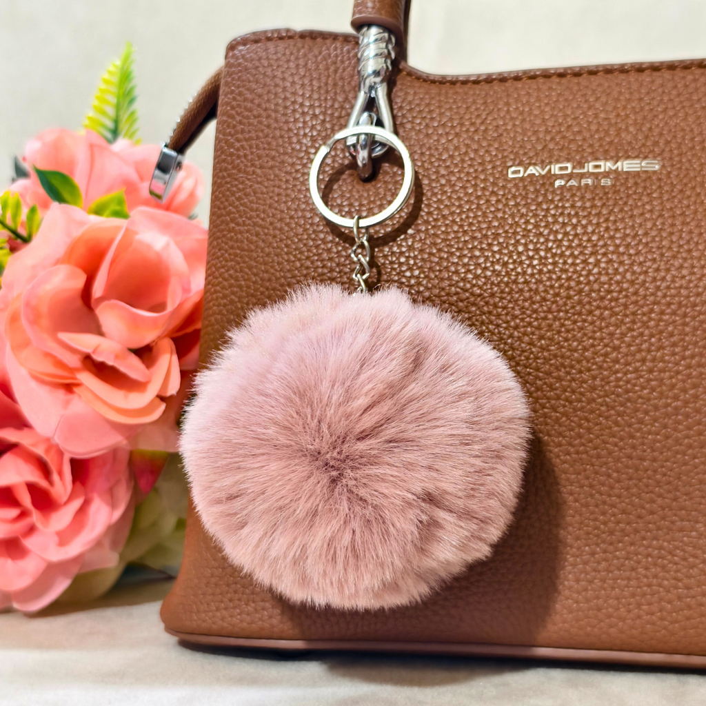 Pink fluffy keychain attached to a brown handbag with flowers in the background